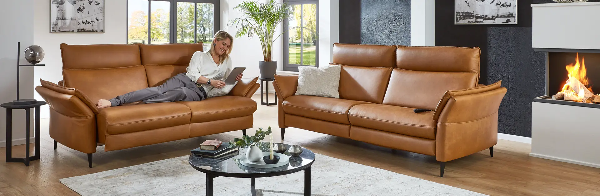 Gemütliches Wohnzimmer mit zwei cognacfarbenen Ledersofas, moderner Einrichtung, Kaminfeuer und entspannter Frau m